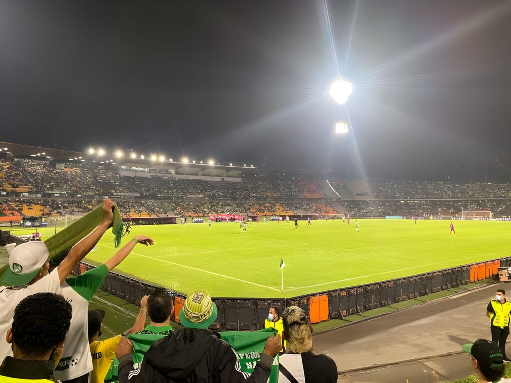 Fans of Atlético Nacional wave flags and cheer during a nighttime match at Estadio Atanasio Girardot, capturing the electric energy of a classic soccer game
